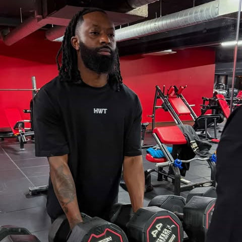 Man holding dumbbells in a gym with red walls and equipment wearing an oversized Tshirt 